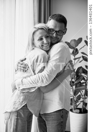 Happy couple hugging on light background at home. Pretty woman smiling and hugging her husband. Sweet couple in casual clothes. Black and white image. Happy couple hugging on light background at home. Pretty woman smiling and hugging her husband. Sweet couple in casual clothes. Black and white image. 136481401