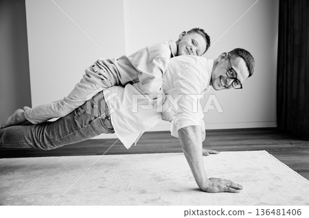 Energetic father and his little son training at home. Kid climbed dad's back while he doing plank. Young family enjoying activity games at home. Black and white image. Energetic father and his little son training at home. Kid climbed dad's back while he doing plank. Young family enjoying activity games at home. Black and white image. 136481406