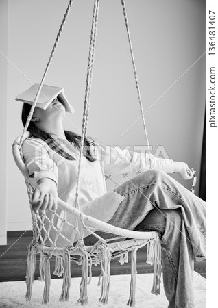 Cute girl covering her face by book. Young female relaxing on indoor hanging chair. Pretty girl in casual clothes chilling on swing with white book on her face. Black and white image. 136481407
