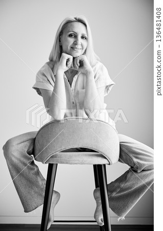 Pretty female relaxing on bar stool. Portrait of woman with blonde hair smiling straight to camera. Beautiful woman sitting on bar stool in her apartment. Black and white image. 136481408