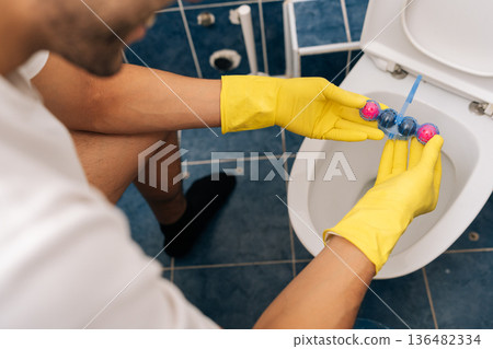 Bearded man in yellow gloves preparing toilet rim block cleaner with gel balls for placement in bathroom toilet for freshness and hygiene. Concept of domestic work, housekeeping and household. Bearded man in yellow gloves preparing toilet rim block cleaner with gel balls for placement in bathroom toilet for freshness and hygiene. Concept of domestic work, housekeeping and household. 136482334