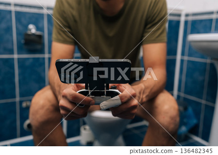 Close-up of unrecognizable man holding smartphone with gamepad attachment and playing video games sitting on toilet, illustrating mobile gaming addiction, multitasking, privacy in bathroom setting. 136482345