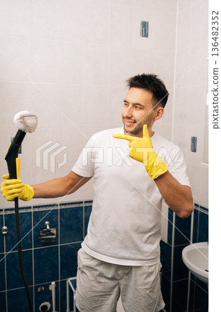 Vertical portrait of smiling man in rubber gloves and white t-shirt using steam cleaner in clean bathroom while pointing finger to appliance, showcasing efficient domestic chores and hygiene. Vertical portrait of smiling man in rubber gloves and white t-shirt using steam cleaner in clean bathroom while pointing finger to appliance, showcasing efficient domestic chores and hygiene. 136482352