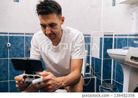 Serious focused young man intently playing mobile video game on smartphone with gamepad, sitting on toilet in bathroom, using technology for leisure and entertainment during daily routine. Serious focused young man intently playing mobile video game on smartphone with gamepad, sitting on toilet in bathroom, using technology for leisure and entertainment during daily routine. 136482356