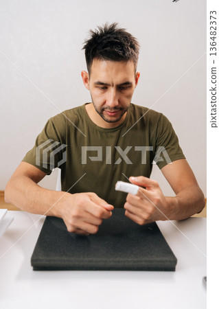 Vertical portrait of young man applying medical patch to injured finger, demonstrating self-care for minor hand wound at home, preventing infection and managing discomfort after accident. Vertical portrait of young man applying medical patch to injured finger, demonstrating self-care for minor hand wound at home, preventing infection and managing discomfort after accident. 136482373