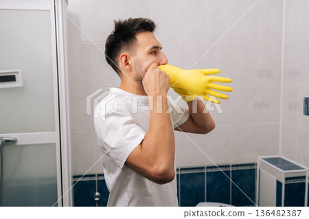 Young man preparing for household chores by putting on yellow protective rubber gloves in bathroom, getting ready for cleaning and sanitation activities. Concept of household chores and hygiene. 136482387
