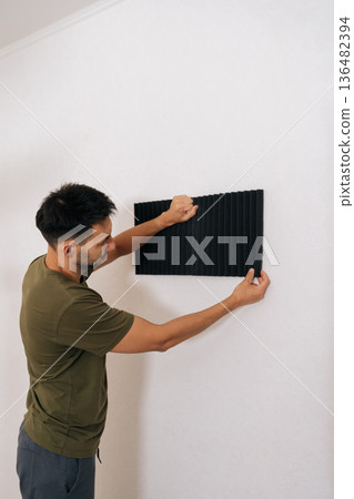 Vertical back view of man fixing applying sound absorbing foam panels to wall, working on acoustic treatment for home studio or office, making soundproofing adjustments and managing noise reduction. 136482394