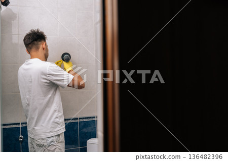 Rear remote view of young man wearing yellow gloves cleaning bathroom tiles using power drill with attached scrubber brush, streamlining household chores and deep cleaning surfaces. Rear remote view of young man wearing yellow gloves cleaning bathroom tiles using power drill with attached scrubber brush, streamlining household chores and deep cleaning surfaces. 136482396