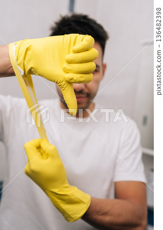 Vertical shot of man holding torn yellow protective glove, making thumbs down gesture to express disappointment, failure or negative feedback related to cleaning tasks or defective equipment. Vertical shot of man holding torn yellow protective glove, making thumbs down gesture to express disappointment, failure or negative feedback related to cleaning tasks or defective equipment. 136482398