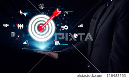 Businessman holding tablet with digital target visualization and various icons representing success, strategy, analytics, and technology in dark backdrop. Qubit Businessman holding tablet with digital target visualization and various icons representing success, strategy, analytics, and technology in dark backdrop. Qubit 136482563