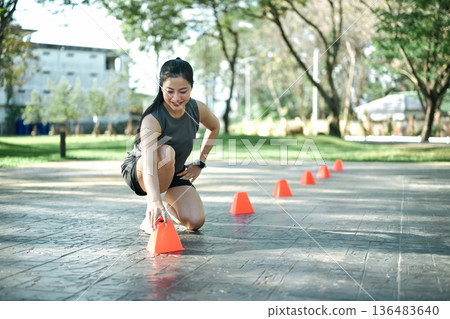 Active woman placing training cones on pavement to prepare agility and speed workout in a public park, Active woman placing training cones on pavement to prepare agility and speed workout in a public park, 136483640