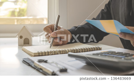 Close-up of a businessman or real estate agent calculating a financial budget and taking notes with a house model on the desk. Mortgage loan application, property investment, and financial planning Close-up of a businessman or real estate agent calculating a financial budget and taking notes with a house model on the desk. Mortgage loan application, property investment, and financial planning 136483669