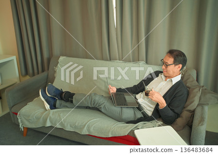 Middle-aged man relaxing on a sofa using a laptop and drinking coffee at home, wearing a covered prosthetic leg, calm independent lifestyle Middle-aged man relaxing on a sofa using a laptop and drinking coffee at home, wearing a covered prosthetic leg, calm independent lifestyle 136483684