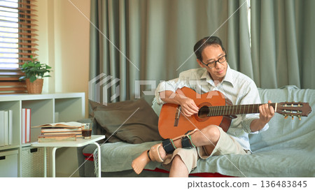 Man playing acoustic guitar on a sofa at home, wearing a prosthetic leg, relaxed leisure time and independent adaptive lifestyle. Man playing acoustic guitar on a sofa at home, wearing a prosthetic leg, relaxed leisure time and independent adaptive lifestyle. 136483845
