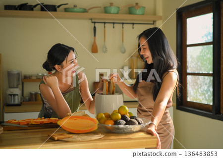 Two young women enjoying a cheerful cooking moment together in a sunlit home kitchen, preparing toast and fresh fruit with laughter Two young women enjoying a cheerful cooking moment together in a sunlit home kitchen, preparing toast and fresh fruit with laughter 136483853