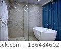 Large oval bathtub in the bathroom. There are two white coats hanging on the wall. Fresh, clean robes. Glass partition in the bathroom interior. The concept of cleanliness and comfort in the room. 136484602