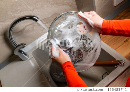 A close-up with localized lighting. The hands of a Caucasian woman washing dishes in a home kitchen late at night in the dark. 136485741