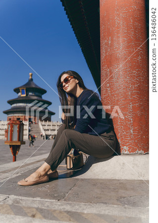 brunette girl sitting on the steps by temple of heaven in China. Brunette girl enjoying chinese architecture while sitting on the steps and relaxing. Caucasian tourist taking a break from exploring brunette girl sitting on the steps by temple of heaven in China. Brunette girl enjoying chinese architecture while sitting on the steps and relaxing. Caucasian tourist taking a break from exploring 136486042