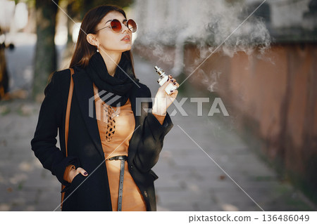 stylish girl smoking an e-cigarette as she is walking through the city stylish girl smoking an e-cigarette as she is walking through the city 136486049