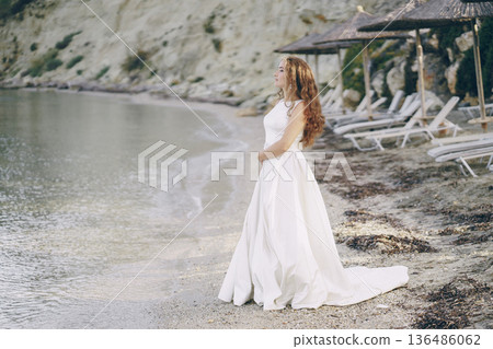 Beautiful long-haired bride in a magnificent white dress walking on a beach Beautiful long-haired bride in a magnificent white dress walking on a beach 136486062