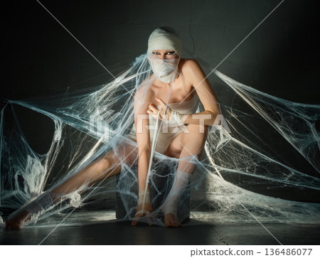 Woman in mummy costume with dramatic eye makeup sitting trapped in artificial spiderwebs in a dark, spooky setting for Halloween. 136486077