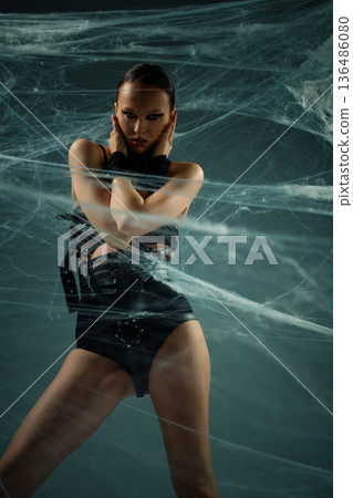 Intense woman in black gothic fashion outfit posing behind a dense spiderweb. Intense woman in black gothic fashion outfit posing behind a dense spiderweb. 136486080