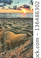Heart shape drawn in sand during sunset at the beach by the ocean with waves and clouds in the background 136486302