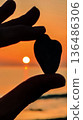 Holding a heart-shaped stone against the backdrop of a sunset over the water creating a moment of connection and reflection 136486306