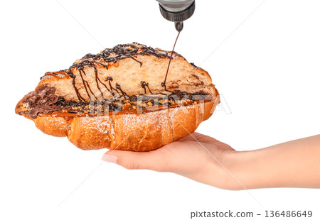 Chocolate syrup pouring over stuffed croissant with caramel and nuts in female hand isolated on white Chocolate syrup pouring over stuffed croissant with caramel and nuts in female hand isolated on white 136486649