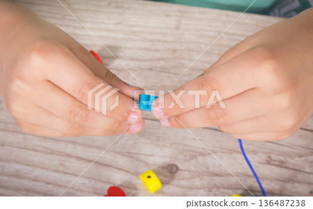 Preschooler making bracelet from thread and wooden colored beads. Development of kids motor skills Preschooler making bracelet from thread and wooden colored beads. Development of kids motor skills 136487238