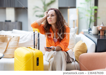 Adult woman sitting with yellow suitcase and holding smartphone in hotel lobby Adult woman sitting with yellow suitcase and holding smartphone in hotel lobby 136487699