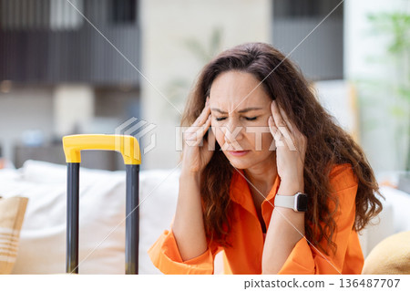 Stressed woman holding her head while sitting near suitcase in hotel lobby Stressed woman holding her head while sitting near suitcase in hotel lobby 136487707