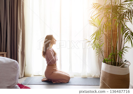 Adult woman practicing yoga kneeling on mat near window in hotel room 136487716