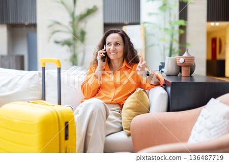 Smiling woman talking on phone while sitting near suitcase in hotel lobby 136487719