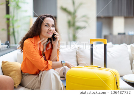Adult woman talking on phone while sitting beside suitcase in hotel lounge Adult woman talking on phone while sitting beside suitcase in hotel lounge 136487722