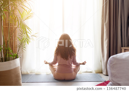 Rear view of woman meditating on yoga mat near window in room Rear view of woman meditating on yoga mat near window in room 136487740