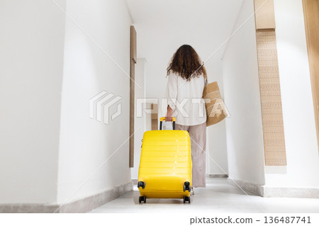 Adult woman walking away through long hotel hallway with suitcase, minimalist interior Adult woman walking away through long hotel hallway with suitcase, minimalist interior 136487741