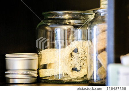 Glass jars with natural bath accessories on a wooden shelf. Loofah sponge, handmade soap and metal cosmetic tins in a cozy bathroom setting Glass jars with natural bath accessories on a wooden shelf. Loofah sponge, handmade soap and metal cosmetic tins in a cozy bathroom setting 136487866