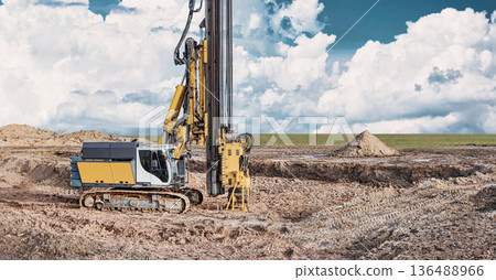 A drilling rig works on a pile foundation at a construction site with dirt and a clear sky filled with clouds in the background A drilling rig works on a pile foundation at a construction site with dirt and a clear sky filled with clouds in the background 136488966