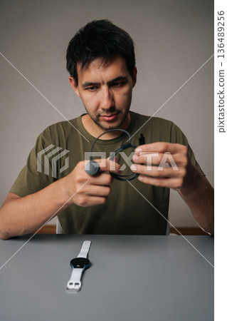 Vertical portrait of pensive young man untangling black charging cable for smart watch sitting at table with watch on isolated background, importance of keeping device powered up. Vertical portrait of pensive young man untangling black charging cable for smart watch sitting at table with watch on isolated background, importance of keeping device powered up. 136489256