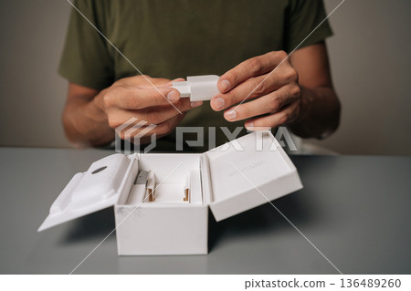 Close-up cropped shot of unrecognizable man unpacking box, holding white smartphone charger being unpacked from cardboard box, sitting on gray table. Concept of contemporary gadget unboxing. Close-up cropped shot of unrecognizable man unpacking box, holding white smartphone charger being unpacked from cardboard box, sitting on gray table. Concept of contemporary gadget unboxing. 136489260