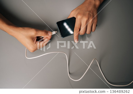 Top view of male hands connecting white usb cable to smartphone on gray surface, charging device, transferring data efficiently. Modern technology facilitating seamless communication and power supply Top view of male hands connecting white usb cable to smartphone on gray surface, charging device, transferring data efficiently. Modern technology facilitating seamless communication and power supply 136489262