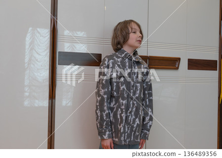 Portrait of young boy standing near wardrobe. Kid wearing casual grey shirt at home 136489356