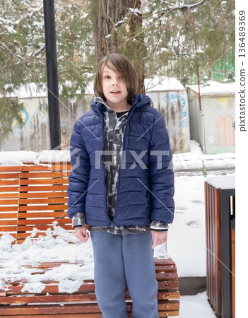 Young boy standing in snowy park. Child in blue winter jacket outdoors. Cold weather lifestyle 136489369