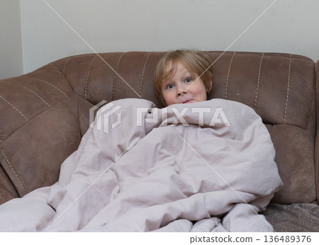 Young boy wrapped in a blanket on a brown sofa. Cute child hiding under a cover at home 136489376