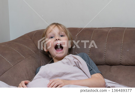 Tired young boy yawning with mouth wide open on a sofa. Sleepy child waking up at home Tired young boy yawning with mouth wide open on a sofa. Sleepy child waking up at home 136489377