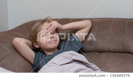 Young boy with a headache touching his forehead on a sofa. Sick child feeling unwell at home Young boy with a headache touching his forehead on a sofa. Sick child feeling unwell at home 136489379