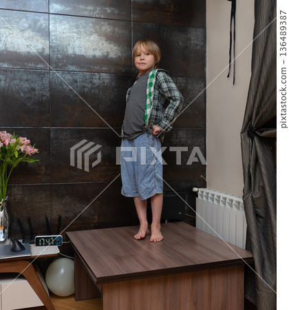 Confident young boy standing on table with hands on hips. Serious child posing on furniture at home 136489387