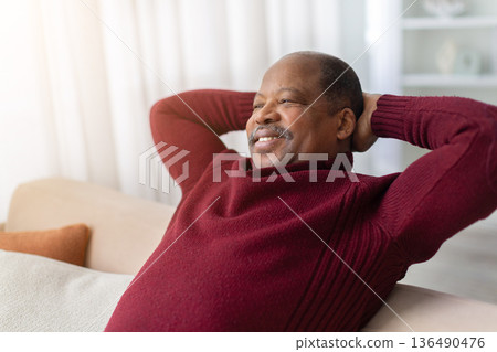 Relaxed black man sitting on sofa at home with hands behind head 136490476