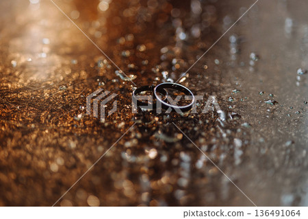 Two wedding rings on wet reflective surface with water droplets and golden bokeh lights 136491064